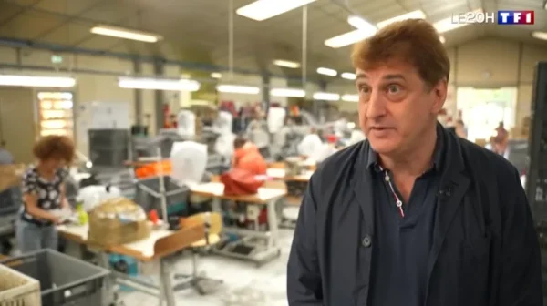 Reportage télévisé TF1 dans l’atelier Airplum : le directeur associé Frédéric Guiral de Haas en chemise sombre s’exprime face caméra, tandis qu’en arrière-plan plusieurs ouvriers travaillent sur des postes de production de chaussons dans un atelier lumineux.