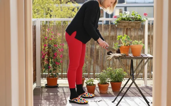 femme blonde aux cheveux mi-longs, vêtue d'un pull noir ample et de leggings rouges, accroupie sur un balcon ensoleillé en train de jardiner. Elle porte des chaussettes noires et des chaussons mules multicolores airplum modèle ondrian, à motifs géométriques (jaune, rouge, bleu, etc.)