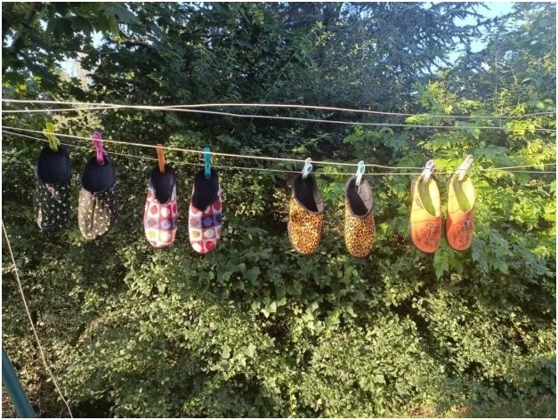 Plusieurs paires de chaussons d'intérieur colorés suspendus avec des pinces à linge sur un fil à linge extérieur dans un jardin verdoyant. Les chaussons présentent différents motifs : imprimés floraux roses, motifs léopard, et couleurs unies (noir, beige, orange, jaune). Séchage naturel en plein air avec végétation dense en arrière-plan